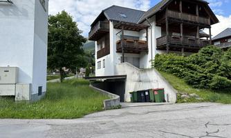 A modern building with balconies stands next to a green meadow. In the foreground, trash bins and a driveway to a garage are visible.