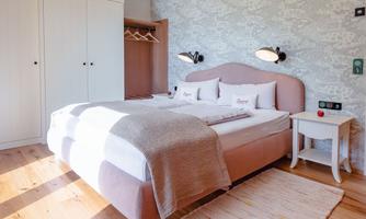A modern bedroom with a large bed and bright wooden flooring. There is a wardrobe and a small nightstand.