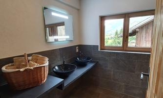 A modern bathroom with wooden shelves and two sinks. The window offers a view of the greenery.