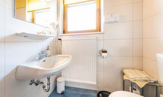 A modern bathroom with a hand wash basin, a toilet, and a large window. The walls are light tiled, and there is pleasant lighting.
