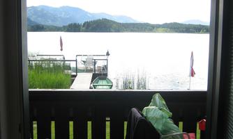 A view from a balcony overlooking a quiet lake with a mountain landscape in the background. In front of the balcony, there are some furniture and a sun umbrella.