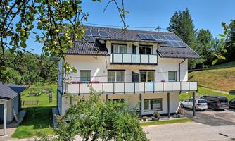 A modern house with solar panels on the roof and a balcony. Surrounded by green spaces and trees.