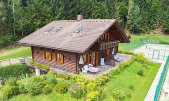 A cozy wooden house surrounded by greenery, with a small patio and a view of a tennis court. The surroundings are peaceful and close to nature.