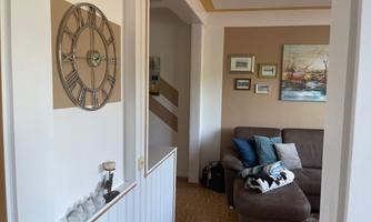 A cozy living space with a brown couch and decorative pictures on the wall. A large wall clock hangs on the wall, and the floor is covered with wooden tiles.