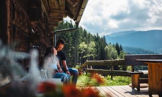 A couple is sitting on a porch in front of a mountain cabin, enjoying the view. In the background, green forests and mountains can be seen.