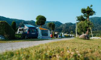 A cozy campsite with caravans and tents in a green landscape. In the background, gentle hills and a clear blue sky can be seen.