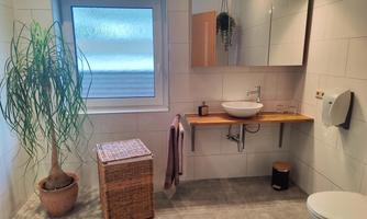 A modern bathroom with a large window, a sink, and a mirror. In the corner stands a plant pot and a laundry basket.