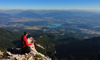 A man is sitting on a rock and looking at a vast landscape. In the background, there are mountains and a clear sky.