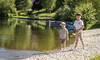 Two boys are standing by the shore of a calm lake. The surroundings are green and sunny, with trees in the background.