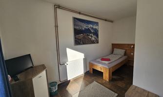A simple room with a single bed, a television, and a wooden desk. On the wall hangs a picture of a mountain landscape.