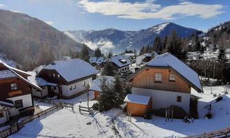 A picturesque winter landscape with snow-covered houses and tall mountains in the background. The sky is clear and blue, creating a serene atmosphere.