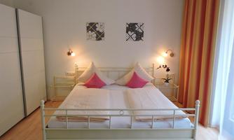 A bright bedroom with a double bed and pink pillows. Two pictures are hanging on the walls, and the room has large windows with curtains.