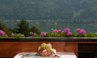 A table with fresh fruit and a magazine is set against a beautiful view of a lake and surrounding flowers. The landscape is characterized by gentle hills and lush greenery.