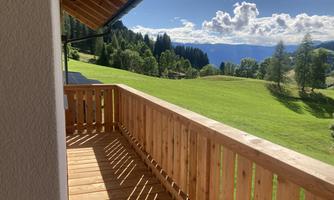 A beautiful balcony with a wooden railing and a view of a green landscape. In the background, there are trees and a blue sky with clouds.