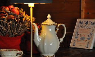 A decorative teapot stands next to a lamp on a table. In the background, a basket of apples and a book about Hygge are visible.