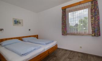 A simple bedroom with a double bed and blue bedcovers. A window with curtains lets in daylight.