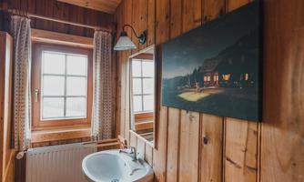 A cozy bathroom with wooden walls and a large window.  
On the wall hangs a picture of a lit house at night.