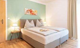 A bright bedroom with a large double bed and two nightstands. A colorful artwork hangs on the wall, and there is a pleasant wooden floor.