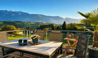 A cozy terrace with a table and chairs, surrounded by a beautiful mountain landscape. The clear sky and the sun give the scene an inviting atmosphere.