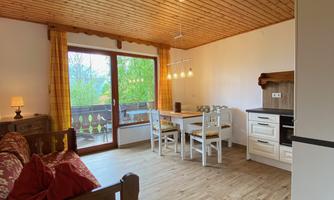 A cozy living room with wooden floors and a wooden ceiling. There is a dining table, chairs, and large windows with a view of the outdoors.