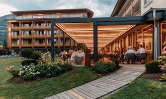A modern restaurant with stylish lighting and many guests outdoors. In the background, an elegant hotel building can be seen.