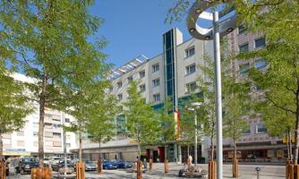 A modern cityscape with a multi-story building and many trees. The street is lively with parked cars and pedestrians.