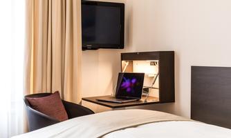A modern hotel room with a bed, a desk, and a television. On the desk is a laptop and there are curtains that dim the light.