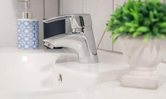 A modern faucet at a sink with a clean design. Next to the faucet is a small plant and a soap dispenser in decorative blue.