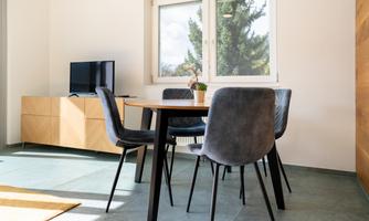 A modern dining room with a table and four chairs. A TV and large windows that let in plenty of daylight are visible.