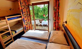 A bright room with bunk beds and a double bed. The windows offer a view of a green surroundings and a small balcony.