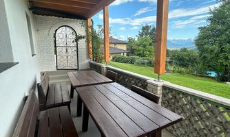 A covered balcony with dark wooden tables and benches. In the background, you can see a green landscape and a blue sky.