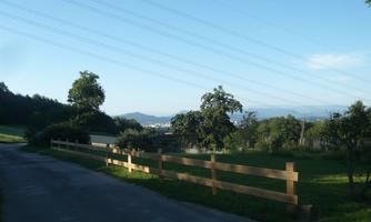 A green landscape with a paved path and a wooden fence. In the background, trees and hills are visible.