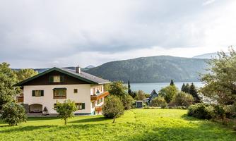 A beautiful house with a large garden and a view of a lake. In the background, the mountains and a cloudy sky are visible.