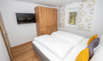 A cozy bedroom with a double bed and bright cushions. A television hangs on the wall, and there is a large wooden wardrobe.