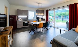 A modern kitchen with a dining area and a cozy living room. Large windows provide a view of the outdoor area.