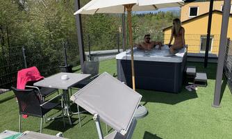 A comfortable outdoor area with a jacuzzi, two people are relaxing in it. There is a sun umbrella, loungers, and a table with chairs.