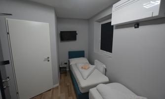 A modern bedroom with two single beds and a television on the wall. The walls are light gray and the floor is made of light wood.