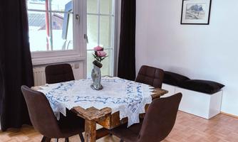 A cozy dining area with a wooden table and four chairs. On the table, there is a vase with a flower, and the windows let in plenty of light.