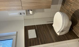 A modern bathroom with a white toilet and wooden elements. The wall is partially tiled, and a window provides natural light.