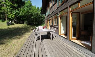 A spacious terrace with a wooden dining area. In the background, green trees and a cozy apartment can be seen.
