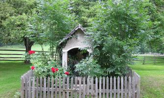 A small, cozy garden house surrounded by green trees and colorful roses. A picturesque fence frames the scene.