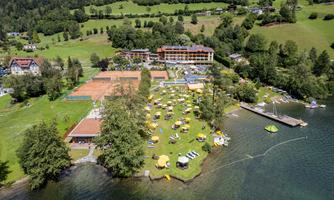 A picturesque landscape by the lake with a well-maintained sunbathing area and numerous sun umbrellas. In the background stands a modern building, surrounded by trees and green meadows.