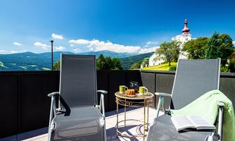 A sunny terrace with two comfortable chairs and a small table. In the background, green hills and a building with a red tower can be seen.