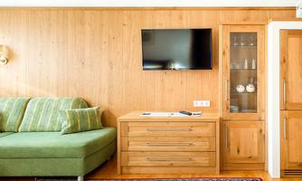 A cozy living room with a green sofa and a wooden table. A television hangs on the wall, next to it is a glass table with dishes.