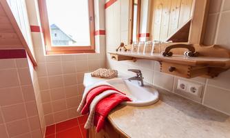 A bright bathroom with a sink and a large window. Towels in red and a wooden shelf complete the cozy atmosphere.