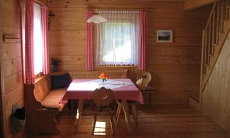 A cozy dining area in a wooden house. The table is set with a pink cloth and there are bright windows with curtains.