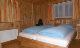 A cozy wooden bedroom with a double bed and blue bedspreads. The walls are clad in wood and the windows are fitted with light curtains.