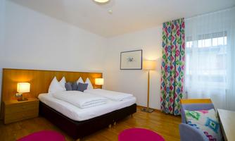 A modern bedroom with a large bed and colorful rugs. The walls are painted bright, and a window provides plenty of daylight.