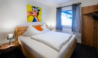 A cozy bedroom with a wooden bed and colorful artwork on the wall. Large windows let in plenty of light and give the room an inviting atmosphere.