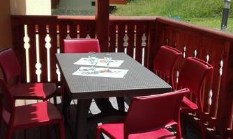A cozy balcony with a black table and red chairs. In the background, some houses and a green meadow can be seen.